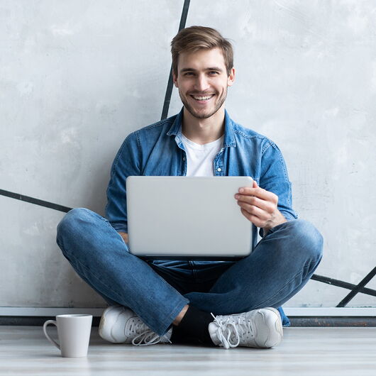 Junger Mann sitzt im Schneidersitz auf dem Boden mit dem Laptop auf dem Schoß und einer Kaffeetasse vor sich.